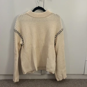 Beige stickad tröja med svarta detaljer - Säljer en beige stickad tröja med svarta dekorativa sömmar längs axlar och ärmar. Tröjan har en högre ribbad krage och långa ärmar med relaxed fit. Perfekt för dig som gillar en enkel men ändå unik look.
