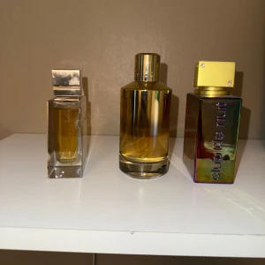 Tre lyxiga parfymer: Amber Oud, Instant Crush, Club de Nuit - Säljer tre exklusiva parfymer: Al Haramain Amber Oud i guldig fyrkantig flaska, Mancera Instant Crush i rund gyllene flaska och Armaf Club de Nuit Untold i regnbågsskimrande fyrkantig flaska med guldfärgat lock och kedja. Alla flaskor har unika och stilrena designer.