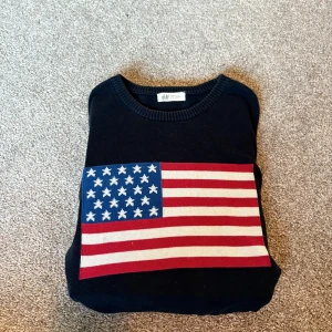 Svart stickad tröja med USA-flagga H&M - Svart stickad tröja från H&M i storlek 170. Framsidan har en stor, färgglad USA-flagga i blått, rött och vitt. Tröjan har rund halsringning och långa ärmar.