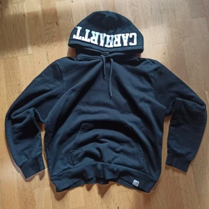 Carhartt hoodie - Svart hoodie från Carhartt med stor vit logga tryckt på huvan. Klassisk känguruficka framtill, dragsko i huvan och ribbade muddar. Perfekt för dig som gillar streetwear och vill ha en stilren look med tydlig branding.10/10 skick. Mer information, bilder och prisdiskutering i dm. Äkta. Passar M