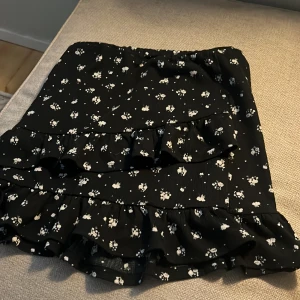 Svart blommig volangkjol Kappahl - Supersöt svart kortkjol från Kappahl med vita små blommor och volangdetaljer. Kjolen har en elastisk midja och är gjord i ett lätt, skönt material💕