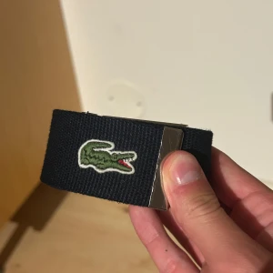 Marinblått Lacoste bälte  - Snyggt blått tygbälte från Lacoste med klassiskt silverfärgat metallspänne där Lacoste-loggan är ingraverad. På bältet finns den ikoniska gröna krokodilen broderad. Perfekt accessoar för att lyfta din outfit med en sportig touch.