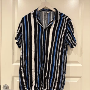 Randig kortärmad skjorta i blått och svart - Snygg skjorta med vertikala ränder i svart, blått och vitt. Skjortan har korta ärmar, klassisk krage och knäppning framtill. Perfekt för dig som gillar en färgstark och trendig look.
