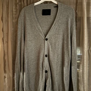 Grå kofta från Jack & Jones - Snygg grå kofta från Jack & Jones med v-ringning och tre knappar framtill. Långärmad och i mjuk bomull, perfekt att slänga över en t-shirt. Enkel och stilren design som funkar till det mesta.