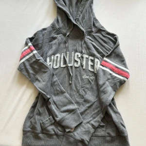 Hollister Hoodie Y2K - Grå hoodie från Hollister med broderat vitt logotyptryck på bröstet och texten 'Los Angeles'. Har huva med snörning, känguruficka och coola ränder i vitt och rött på ärmarna. Mjuk bomullsblandning, perfekt för chill dagar. Y2K