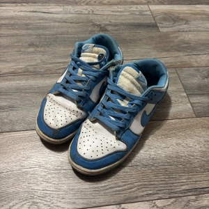 Nike Dunk Low blå och vita sneakers - Nike Dunk Low sneakers i blått och vitt med klassisk swoosh på sidan. Skorna har perforerad tå, snörning och låg profil. Ovandelen är i skinn och innerfodret är blått. Perfekta för dig som gillar retrostil och streetwear.