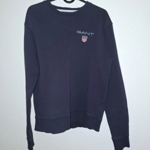 Mörkblå sweatshirt från GANT - Säljer en mörkblå sweatshirt från GANT med broderad logga och sköld på bröstet. Tröjan har rund hals, långa ärmar och ribbade muddar. Perfekt för dig som gillar klassisk och stilren design.