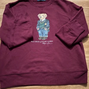 Vinröd Polo Bear tröja Ralph Lauren - Säljer en vinröd sweatshirt från Ralph Lauren med ikoniska Polo Bear-trycket på bröstet. Tröjan har rund hals, långa ärmar och är gjord i mjuk bomull. Perfekt för dig som vill ha en klassisk och snygg look med en twist.