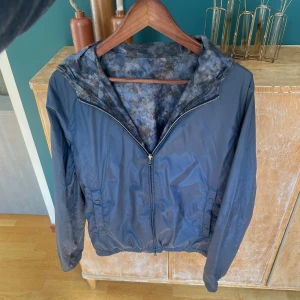 Herno Jacka - Vändbar vindjacka i mörkblått med dragkedja och huva. Ena sidan är enfärgad blå, andra sidan har ett unikt blå-grått mönster. Jackan har resår i midjan och praktiska fickor. Perfekt för dig som vill variera din stil och hålla dig skyddad mot vinden.