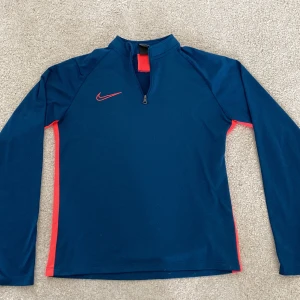 Nike Dri-Fit blå träningshoodie L - Hej! Jag säljer en Nike Dri-Fit långärmad hoodie i marinblått med röda detaljer längs sidorna och axlarna. Tröjan har half zip framtill och är gjord i ett lätt, syntetiskt material som andas. Perfekt för träning! Nike-märket sitter snyggt på bröstet. Den är i storlek L och passar personer mellan 147-158. Perfekt för den som vill kombinera mode med träning!Hör gärna av er vid frågor!