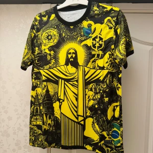 Brasil CBF gul och svart fotbollströja - Unik Brasil CBF fotbollströja med ett stort tryck av Kristusstatyn och detaljerade illustrationer i svart och gul. Tröjan har rund hals, korta ärmar och Brasil-logga på bröstet. Materialet är lätt och andas, perfekt för match eller häng med kompisar.