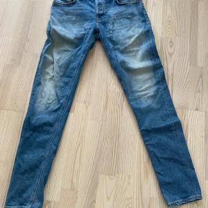 Nudie Jeans Grim Tim blå jeans 30/32 - Säljer ett par Nudie Jeans Grim Tim i klassisk blå tvätt med snygga slitningar och orange sömmar. Modellen är grim tim och sitter slim fitTillverkade i 98% bomull och 2% elastan för lite stretch. Perfekta för dig som gillar en tidlös jeanslook.