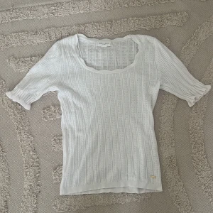 Vit stickad topp från  Holly & Whyte - Supersöt vit stickad topp från Holly & Whyte med korta ärmar och snyggt ribbat mönster. Toppen har en rundad, lite vågig urringning och fina detaljer vid ärmsluten. 