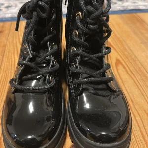 Svarta lackade boots med snörning - Snygga svarta boots i lackad finish med grov sula och svarta snören. Guldiga öljetter ger en extra touch. Skorna har rund tå och platt sula, perfekta för dig som vill sticka ut med en cool och trendig look.