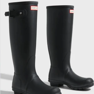 Svarta höga stövlar från Hunter - Klassiska svarta höga stövlar från Hunter med rund tå och justerbart spänne vid skaftet. Tillverkade i slitstarkt gummi med logotyp framtill. Perfekta för regniga dagar och festivaler.