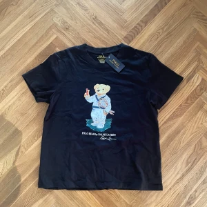 Svart Polo Bear t-shirt från Ralph Lauren - Svart t-shirt från Polo Ralph Lauren med ikoniskt Polo Bear-tryck på bröstet där björnen håller en tennisklubba och dryck. Klassisk rund hals, kort ärm och slim fit. Tillverkad i mjuk bomull för en skön känsla.