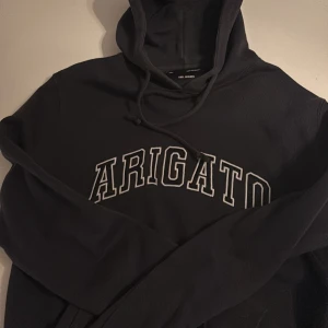 Mörkgrå hoodie Axel Arigato - Mörkgrå hoodie från Axel Arigato med stor vit broderad text på bröstet. Klassisk modell med huva och snörning, samt långa ärmar. Perfekt för en chill och stilren look. Materialet är mjukt och känns skönt mot huden. 