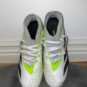 Adidas Predator vita och neon fotbollsskor - Snygga Adidas Predator fotbollsskor i vitt med neon-gula och svarta detaljer. Skorna har snörning, grå plös och hög definition grip-textur för extra bollkontroll. Sulan är grå och neon-gul med fasta dobbar, perfekt för spel på gräs.