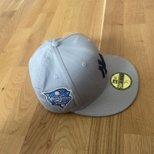 Grå New Era Yankees keps 59Fifty - Snygg grå New Era 59Fifty keps med mörkblå Yankees-logga framtill och World Series 2009 patch på sidan. Platt skärm och klassisk passform, tillverkad i bomull. Perfekt för dig som gillar baseball och streetwear.