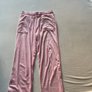 Rosa velourbyxor från Juicy Couture - Mysiga rosa mjukisbyxor från Juicy Couture i velour med bred resår och snörning i midjan. Byxorna har stora fickor fram och bak, varav bakfickorna har lock med tryckknappar. Perfekta för en chill och trendig look.