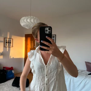 Vit ärmlös linneskjorta med volang - Snygg vit blus i linne med knappar framtill och volangdetaljer längs axlarna. Bluseb är ärmlös och har en avslappnad passform med klassisk krage. Perfekt för varma dagar och ger en fräsch och stilren look.