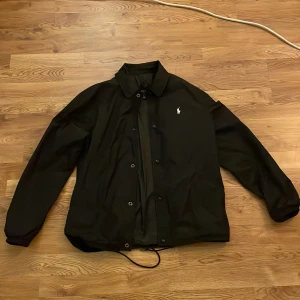 Svart vindjacka Polo Ralph Lauren  - Tja säljer mina svarta vindjacka från Polo Ralph Lauren med klassisk krage, knäppning framtill och vit broderad logga på bröstet. Jackan har dragsko i nederkant och elastiska muddar vid ärmslut. Perfekt för dig som gillar stilren och enkel outfit. Inga defekter!
