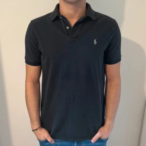 Svart Polo Ralph Lauren Pike - Svart Pike från Polo Ralph Lauren med korta ärmar och krage. Snyggt broderad logga på bröstet och två knappar vid halsen. Modellen är Classic Fit och materialet är mjuk bomull som känns skön mot huden. Perfekt för en chill och stilren look. Mycket bra skick 