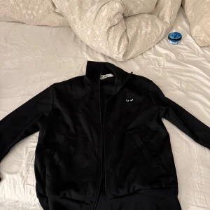 Svart jacka Comme des Garçons Play S - Svart jacka från Comme des Garçons Play med dragkedja och lång ärm. Ikoniskt hjärta med ögon på bröstet och stort hjärta med ögon på ryggen. Snygg och stilren design, perfekt för dig som gillar streetwear och vill sticka ut.