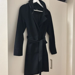 Svart kappa med bälte från BIKBOK  - Stilren svart kappa från BIKBOK med klassisk krage och knytskärp i midjan. Långärmad modell med två fickor framtill och snyggt fall. Perfekt för dig som gillar en clean och tidlös look.