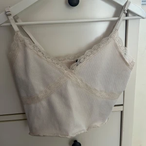 Beige ribbad linnetopp med spets - Supersöt beige linnetopp med tunna axelband och ribbad struktur. Toppen har fina spetsdetaljer längs urringningen och under bysten, vilket ger en romantisk vibe. Perfekt att styla med jeans eller kjol för en trendig look.