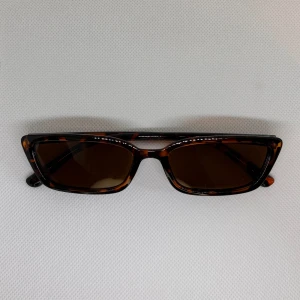 Solglasögon  - Trendiga solglasögon med smal, rektangulär form och bruntonade glas. Bågarna har ett snyggt leopardmönster i brunt och svart som ger en cool retrovibe. Perfekta för dig som vill sticka ut med en unik accessoar till sommaren.