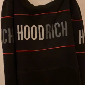 Svart HOODRICH hoodie XL med röd detalj - Svart hoodie från HOODRICH i storlek XL för tonåringar. Tröjan har stora HOODRICH-tryck i grått på bröstet och ärmarna, samt tunna röda linjer som ger en snygg kontrast. Klassisk huva och långärmad modell, perfekt för streetwear-stilen.