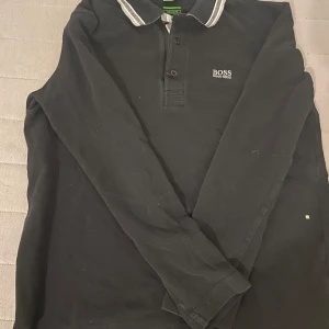 Svart långärmad pikétröja Hugo Boss - Svart långärmad pikétröja från Hugo Boss med vit broderad logga på bröstet. Kragen har vita och grå ränder och tröjan har knappar framtill. Materialet är bomull och passformen är normal. Perfekt för dig som gillar stilrena och klassiska plagg. Regular fit som S