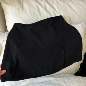 Svart kortkjol med slits - Säljer en svart kortkjol med stilren design och en snygg slits framtill. Kjolen har en figurnära passform och är perfekt att matcha med boots eller klackar. Enkel och trendig look som passar till många olika outfits.