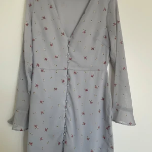 Ljusblå blommig klänning från NLY Trend - Supersöt ljusblå klänning från NLY Trend med små blommor i rött och vitt. Klänningen har v-ringning, långa ärmar och knappar framtill. Den är kort i modellen och har en flowy känsla i tyget, perfekt för vår och sommar.