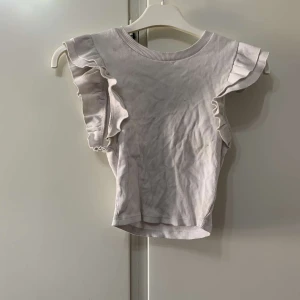 Ljusribbad topp med volangärmar - Supersöt ljus topp i ribbad bomull med volangdetaljer på axlarna. Rund halsringning och ärmlös design som ger en fräsch och trendig look. Perfekt att matcha med jeans eller kjol för en chill stil.