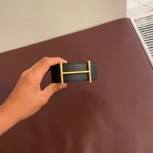 Svart och orange Hermees skärp i skinn - Säljer ett snyggt Hermees bälte i skinn med en bred design och ikonisk H-spänne i guld. Bältet är vändbart med svart på ena sidan och orange på andra, vilket gör det superflexibelt för olika outfits. Perfekt accessoar för att lyfta din stil.