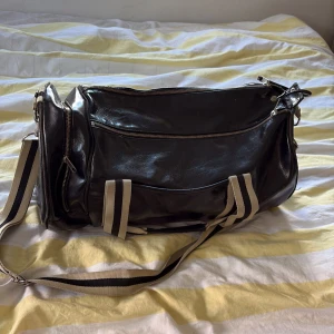 Svart Fred Perry weekendbag i skinn - Snygg svart weekendbag från Fred Perry i glansigt skinn med beige och svarta ränder på axelremmen. Väskan har rundade kortsidor, dragkedja upptill och extra fack på sidan. Perfekt för gymmet eller en kort resa.       Ny pris 1500