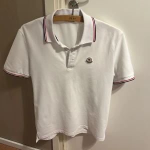 Vit Moncler pikétröja med röd/blå detaljer - Snygg vit pikétröja från Moncler i bomull med klassisk krage och korta ärmar. Tröjan har broderad Moncler-logga på bröstet och stilrena rödblå ränder på krage och ärmslut. Perfekt för dig som gillar clean och sportig stil. Passar även som S.