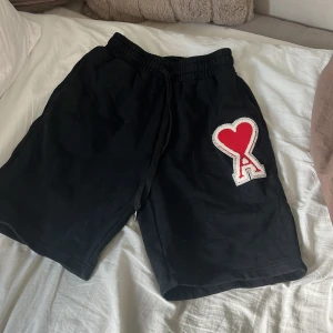 Ami shorts - Svarta shorts från Ami med elastisk midja och snörning. På vänster ben finns en stor röd hjärtdetalj med ett vitt A. Materialet är mjuk bomull, perfekt för chill dagar. Passformen är loose och längden går till knäna.