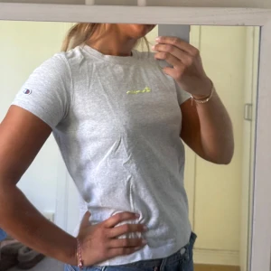 Grå t-shirt Champion - Snygg grå t-shirt från Champion i storlek small. T-shirten har korta ärmar, rund hals och ett litet Champion-logo broderat på ärmen samt tryckt i gult på bröstet. Mjuk och skön bomullskvalitet, perfekt till jeans eller shorts.