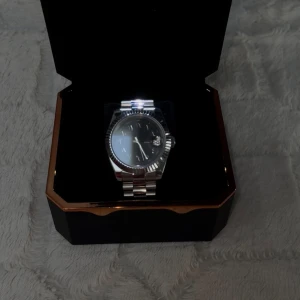 Silverfärgad Seiko klocka med svart urtavla - Stilren Seiko klocka i silverfärgat rostfritt stål med svart urtavla och datumvisning. Klassisk rund form, räfflad bezel och länkarmband ger en tidlös look. Perfekt accessoar för dig som gillar en clean och modern stil.