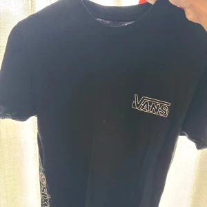 Svart Vans t-shirt med tryck - Svart t-shirt från Vans med klassisk logga på bröstet och stort tryck på ryggen. T-shirten har rund hals och korta ärmar, samt tryck längs ena sidan. Perfekt för dig som gillar streetwear.