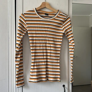 Randig långärmad tröja Gina Tricot - Supersnygg långärmad tröja från Gina Tricot i storlek XS. Tröjan har ett randigt mönster i vitt, svart och orange med ribbad struktur. Passformen är slim och materialet känns mjukt och stretchigt, perfekt till jeans eller kjol. Rund halsringning och smala ärmar.