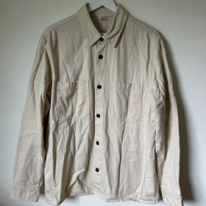 Beige overshirt från Arket, strl 52 - Snygg beige overshirt från Arket i storlek 52. Tillverkad i kraftig bomull med två stora bröstfickor och svarta knappar framtill. Klassisk krage och långa ärmar med knappslå. Perfekt för lager-på-lager och enkel att styla till jeans eller chinos. 1F5