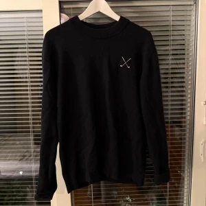 Svart stickad tröja J.Lindeberg M - Svart stickad tröja från J.Lindeberg i storlek M. Framsidan har en liten broderad logga med två korsade golfklubbor och baksidan pryds av en stor vit blixt. Tröjan har rund hals och långa ärmar, perfekt för dig som gillar stilrena detaljer med en twist.