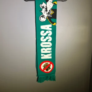 Hammarby halsduk  - Supporterhalsduk i grön färg med vit text 'KROSSA GNAGET' och illustration av en råtta i supporterkläder. Halsduken har fransar i ändarna och ett tryck med överstruket AIK-märke. Perfekt för dig som vill visa ditt stöd på läktaren.