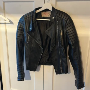 Svart skinnjacka från Voyelles - Snygg svart skinnjacka från Voyelles i bikerstil med markerade axlar och quiltade detaljer på ärmarna. Jackan har dragkedjor både framtill och vid ärmsluten, samt coola silvriga knappar. Perfekt för dig som vill ha en edgy look.