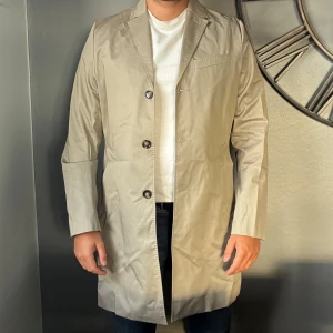 Trenchcoat J.Lindeberg (oanvänd) - Stilren beige rock från J.Lindeberg med klassisk krage, tre knappar framtill och raka fickor. Helt oanvänd, finns fortfarande prislapp på (ord. 2800kr)