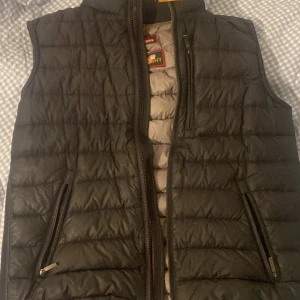 Svart quiltad väst från Parajumpers (äkta) - Säljer en svart quiltad väst från Parajumpers med dragkedja framtill och två praktiska fickor. Västen har en stilren design och är perfekt för kyligare dagar. Den har en diskret logga på baksidan och en bekväm passform. Dock en defekt på västen vid framsidan som verkligen inte syns
